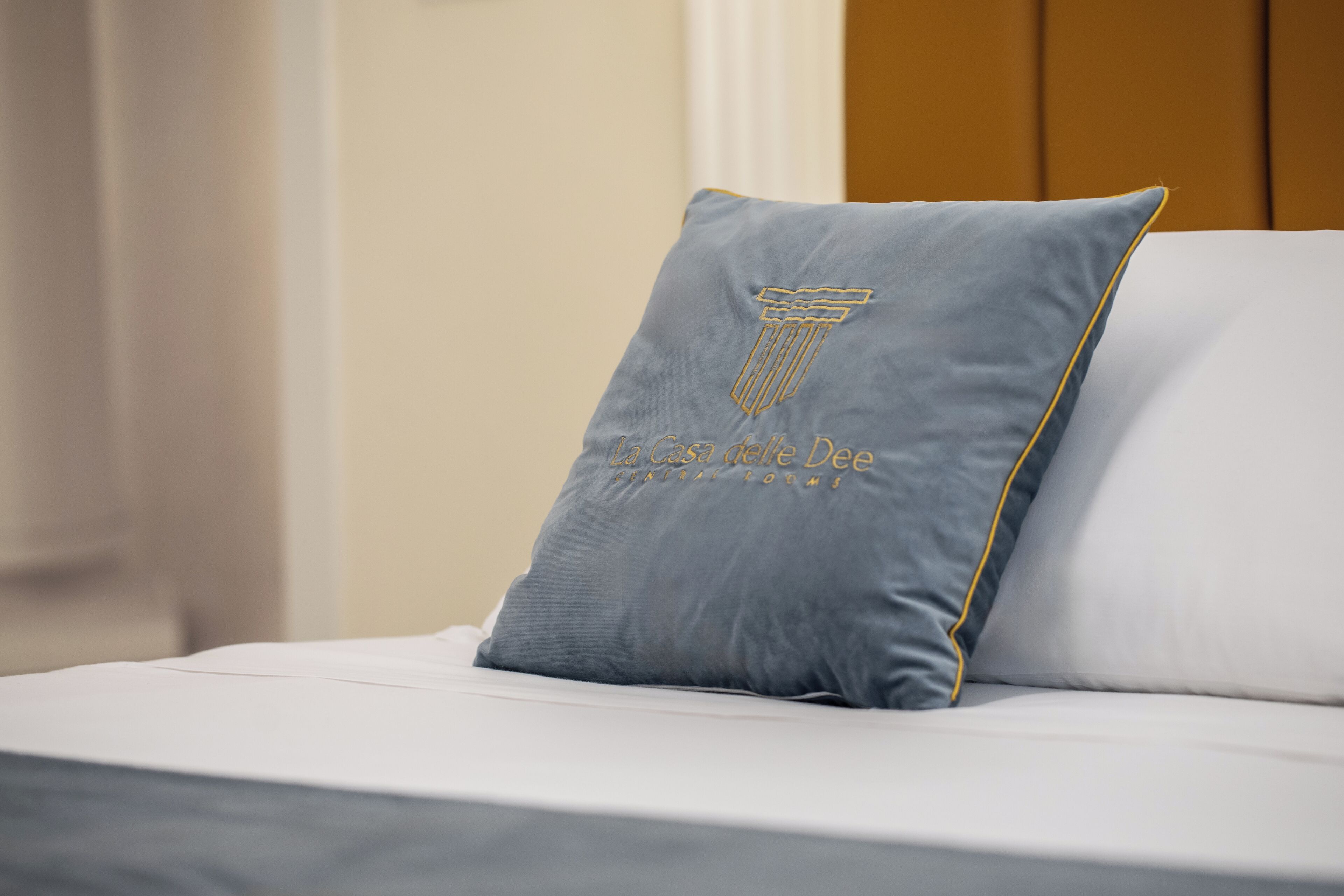 A bed with a grey pillow featuring a column design and the text "La Casa delle Dee" on a white bedspread.