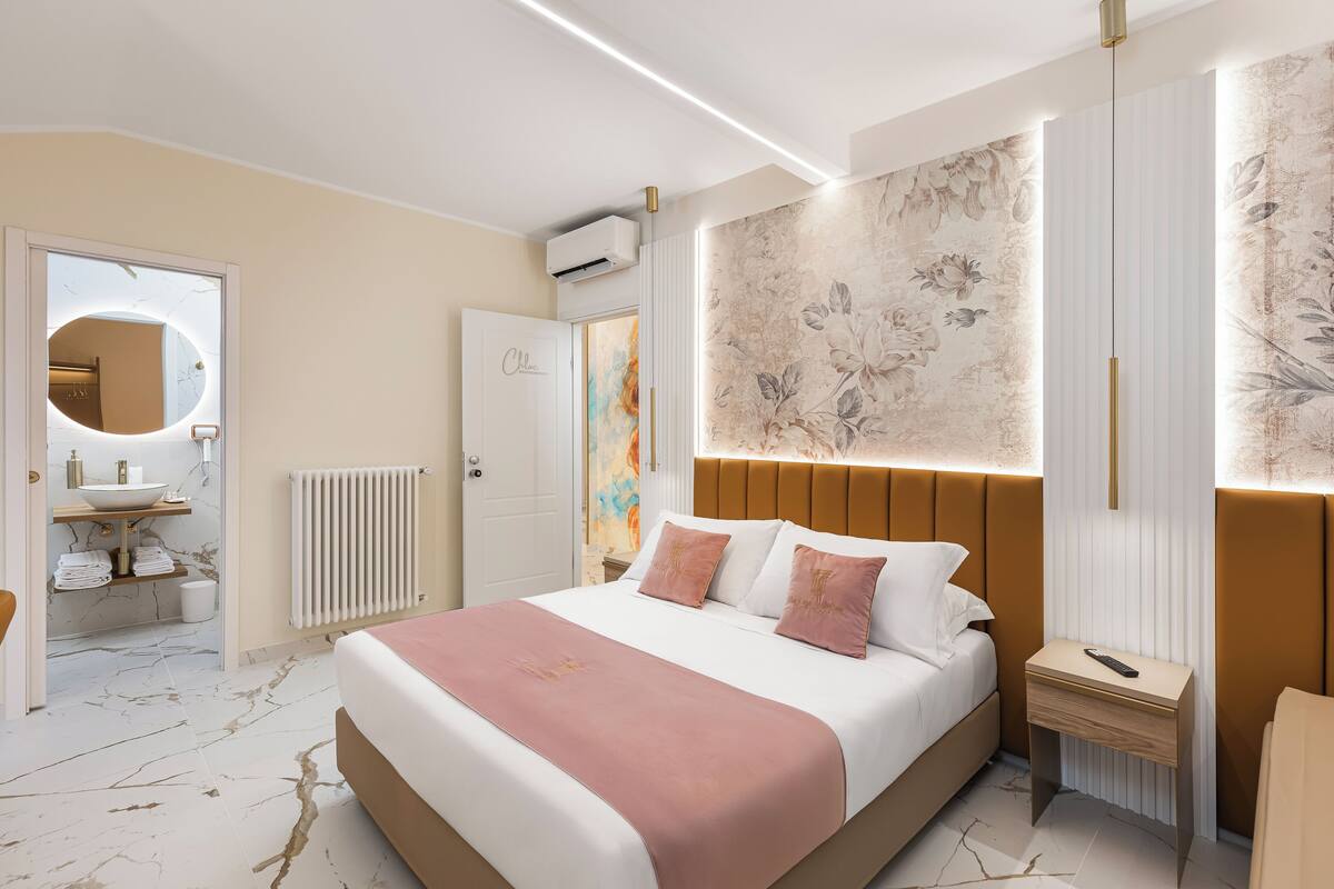 Ein modernes Schlafzimmer mit einem großen Bett, Nachttischen, einem Spiegel und einem Badezimmer mit Waschbecken und Handtuchhalter.