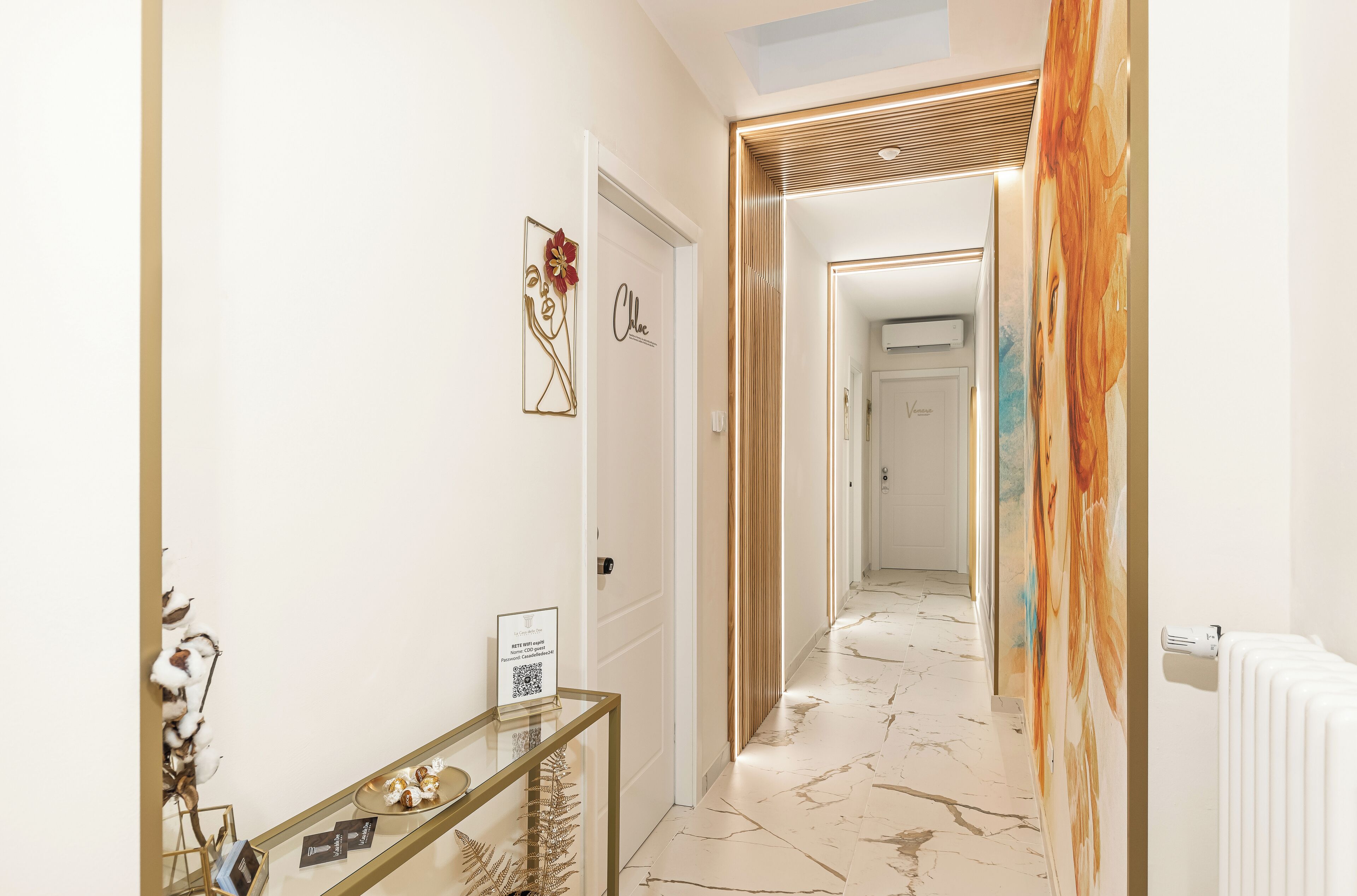 A hotel hallway with a modern design, featuring a glass console table with decorative items, a door with a nameplate, and a corridor leading to another room.