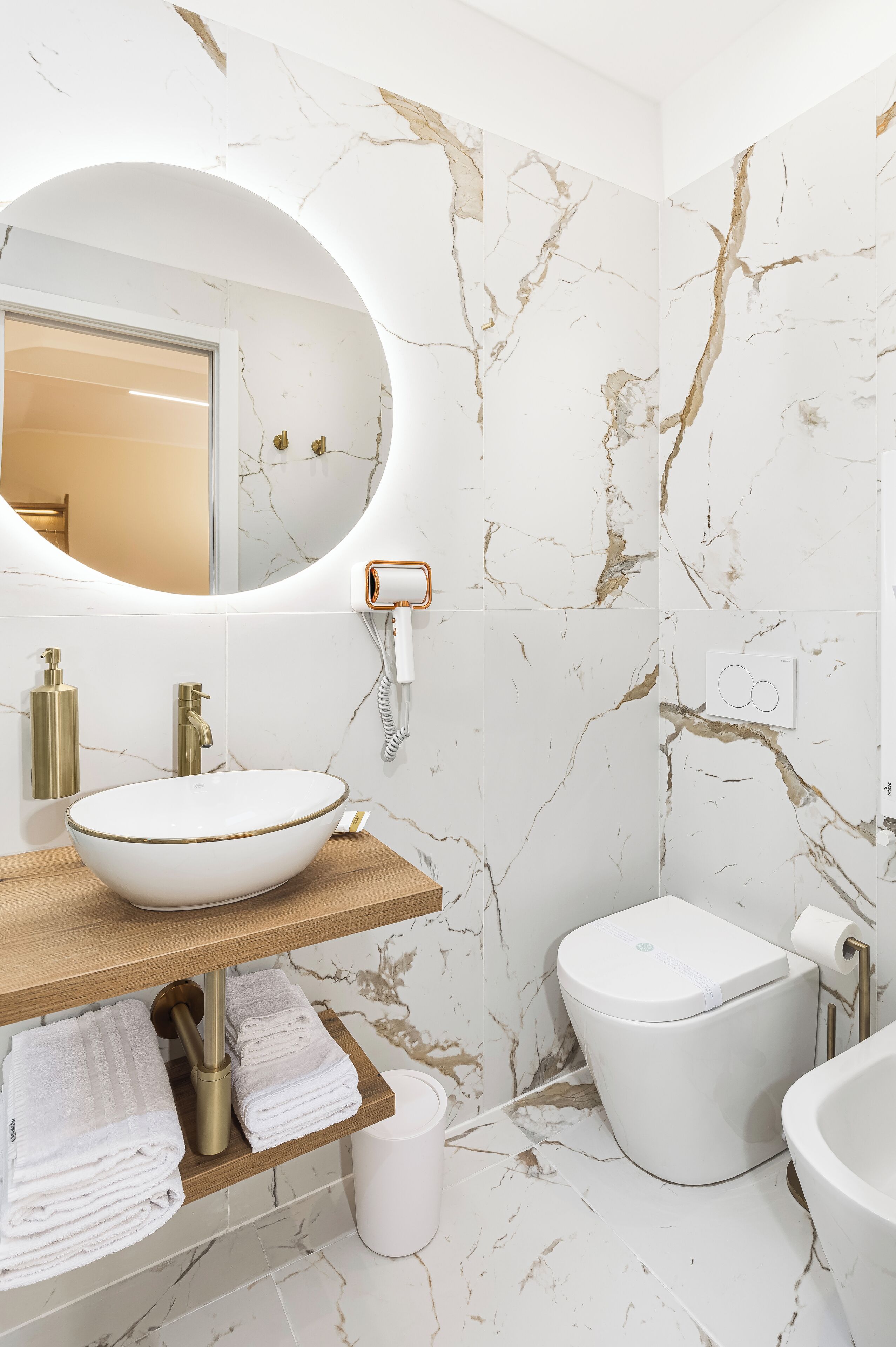 Un baño con pared de mármol, espejo redondo, lavamanos blanco y un inodoro.