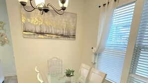 A dining area with a glass-top table, white chairs, a chandelier, a painting on the wall, and a window with blinds. - The Tigers Pathway House near LSU Stadium - BC1034 (Baton Rouge)