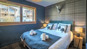 Een net opgemaakt bed met een blauwe deken en twee handdoeken, een raam met uitzicht op gebouwen, een aan de muur bevestigd decoratie van een hertenhoofd en een bedlampje.