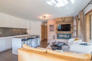 A modern kitchen with white cabinets, a central island, and a dining area with bar stools. There is a living room with a sofa and a fireplace. - Appartement Prariand - Welkeys (Megève)