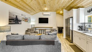 Ein modernes Wohnzimmer mit grauem Sofa, Holzboden und einer Küchenzeile mit weißen Schränken und einem Edelstahlkühlschrank.