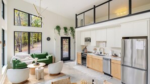 Eine moderne Küche mit Edelstahlgeräten, eine Essecke mit Holztisch und -stühlen sowie ein Wohnbereich mit einem grünen Sofa und weißen Sesseln.