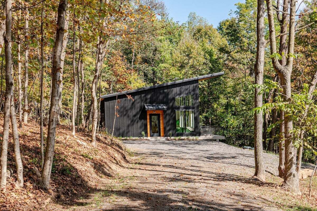 A modern cabin with a dark exterior, a wooden door, and large windows, surrounded by a forested area.