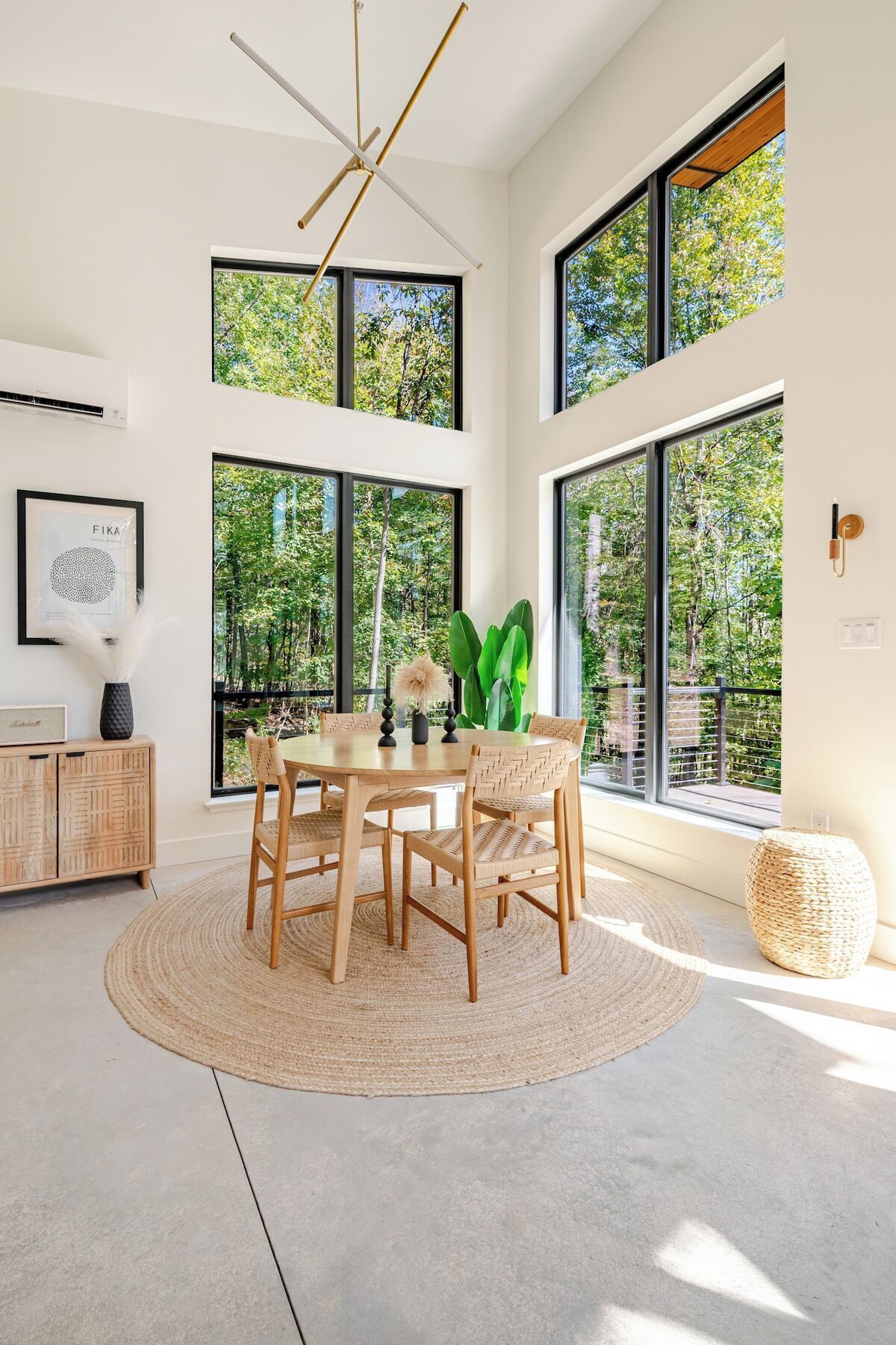 Un espace repas moderne avec une table ronde, des chaises en osier et une grande fenêtre donnant sur un paysage verdoyant.