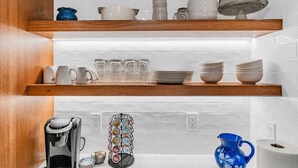 Une cuisine avec des murs recouverts de carreaux blancs, des étagères en bois et divers ustensiles de cuisine, notamment des tasses, des assiettes et une machine à café.