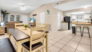 Un grand espace de vie comprenant une table à manger, des chaises, un canapé, un ventilateur de plafond et un coin cuisine équipé d’un réfrigérateur et de tabourets de bar.