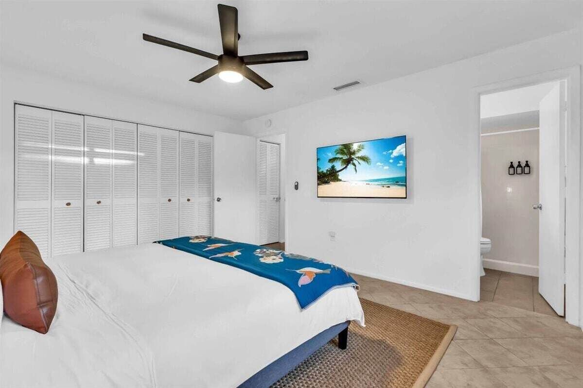 Un dormitorio con una cama, un ventilador de techo, un adorno de pared con motivos playeros y un baño visible a través de una puerta abierta.