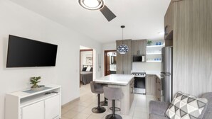 A modern kitchen with a central island, bar stools, a flat-screen TV, and a dining area with a sofa. - Lovely modern 2 bedroom gated condo, walk to the beach and town center (Playas del Coco)