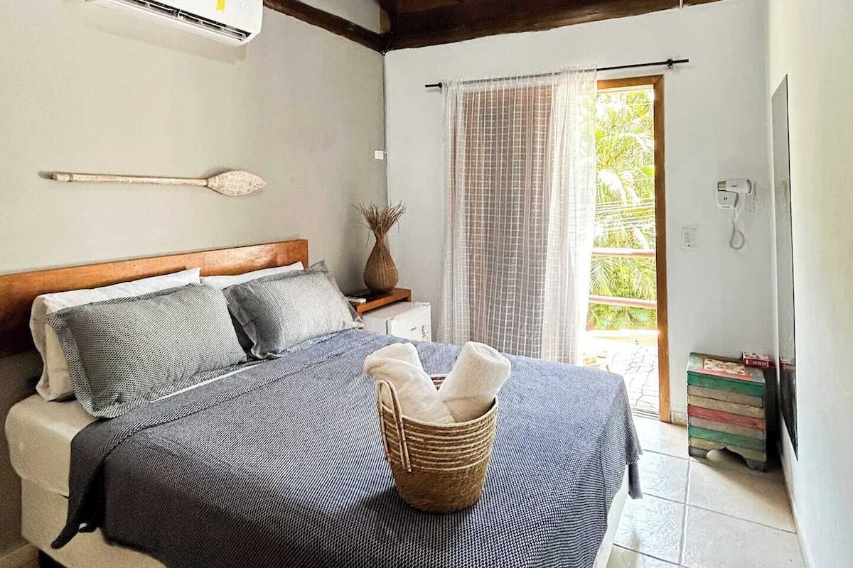A bedroom with a bed, a wall-mounted air conditioner, a wooden ceiling, a window with curtains, and a small bedside table with a vase.