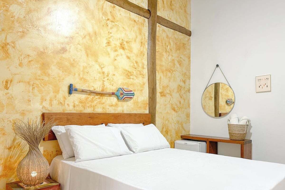 A bedroom with a wooden headboard, white bedding, a bedside table with a lamp, a wall-mounted mirror, and a small basket.