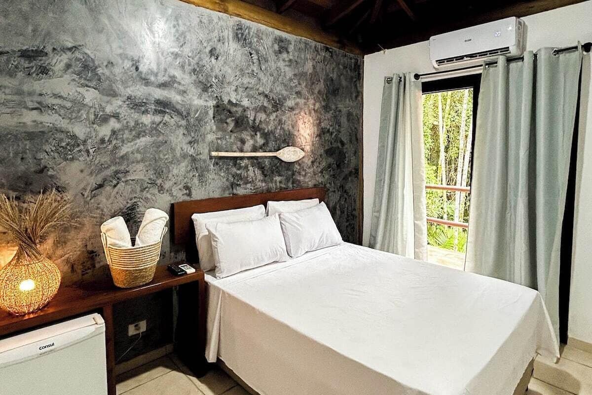 A bedroom with a bed, nightstands, a small refrigerator, a ceiling fan, and a window with curtains.