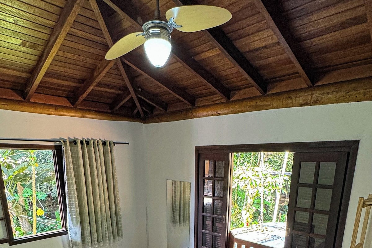 A spacious room with a large bed, a ceiling fan, and a view of greenery outside.