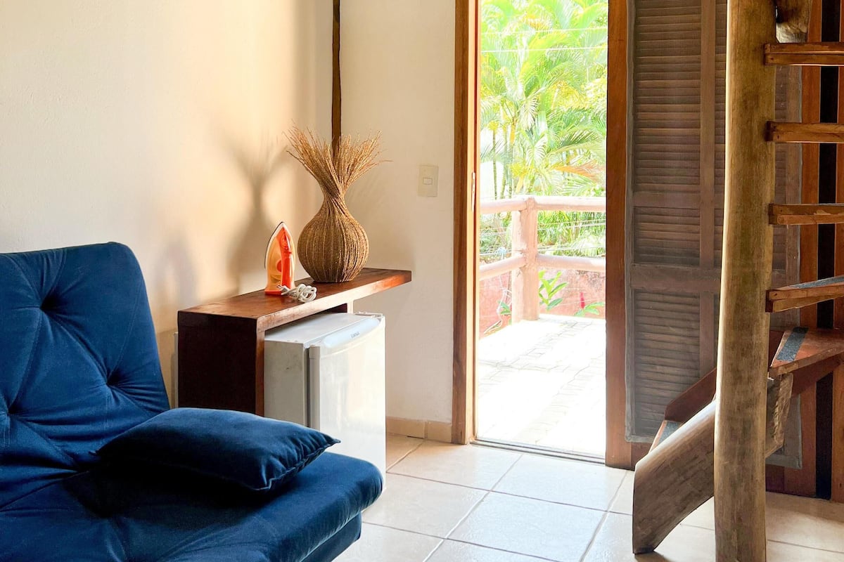 A room with a blue sofa, a small table with a vase, and a wooden staircase leading to a balcony with a view of palm trees.