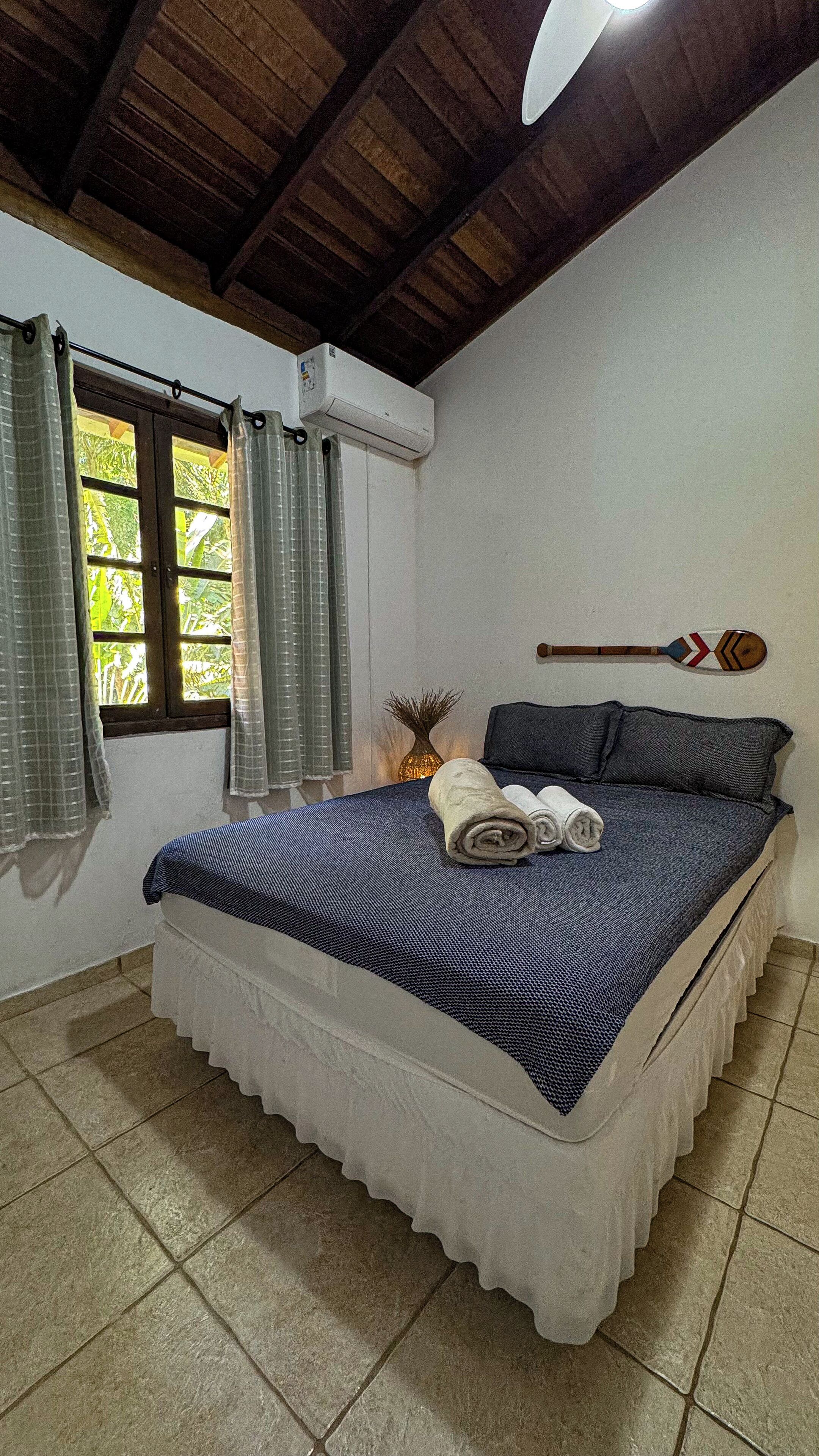 A bedroom with a bed, curtains, an air conditioning unit, a wooden paddle, and a vase.