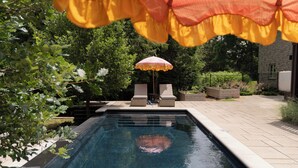 A swimming pool with two lounge chairs and a small table, surrounded by greenery and a stone building.