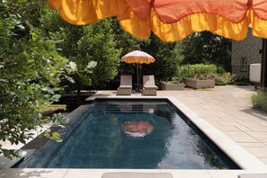 A swimming pool with two lounge chairs and a small table, surrounded by greenery and a stone building.