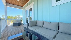 A covered porch with a cushioned seating area, a glass table, and a view of the surrounding landscape.