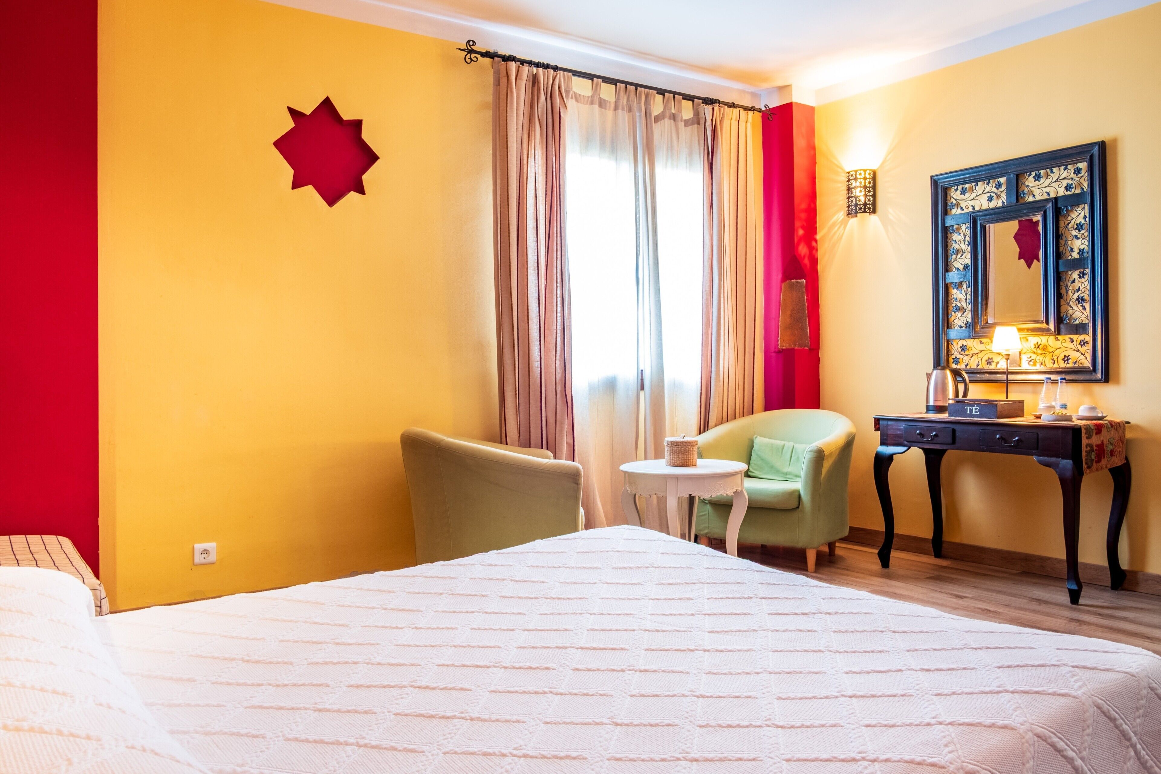 A hotel room with a bed, a desk, a chair, a small table, and a wall-mounted lamp.