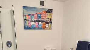 Une salle de bain avec une baignoire, des toilettes et un tableau représentant des maisons colorées accroché au mur.