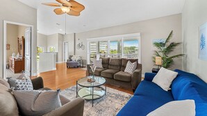 Ein geräumiges Wohnzimmer mit einem blauen Sofa, einem braunen Sofa und einem Glastisch. Es gibt einen Deckenventilator, ein Fenster mit Jalousien und eine Pflanze auf einem Beistelltisch.