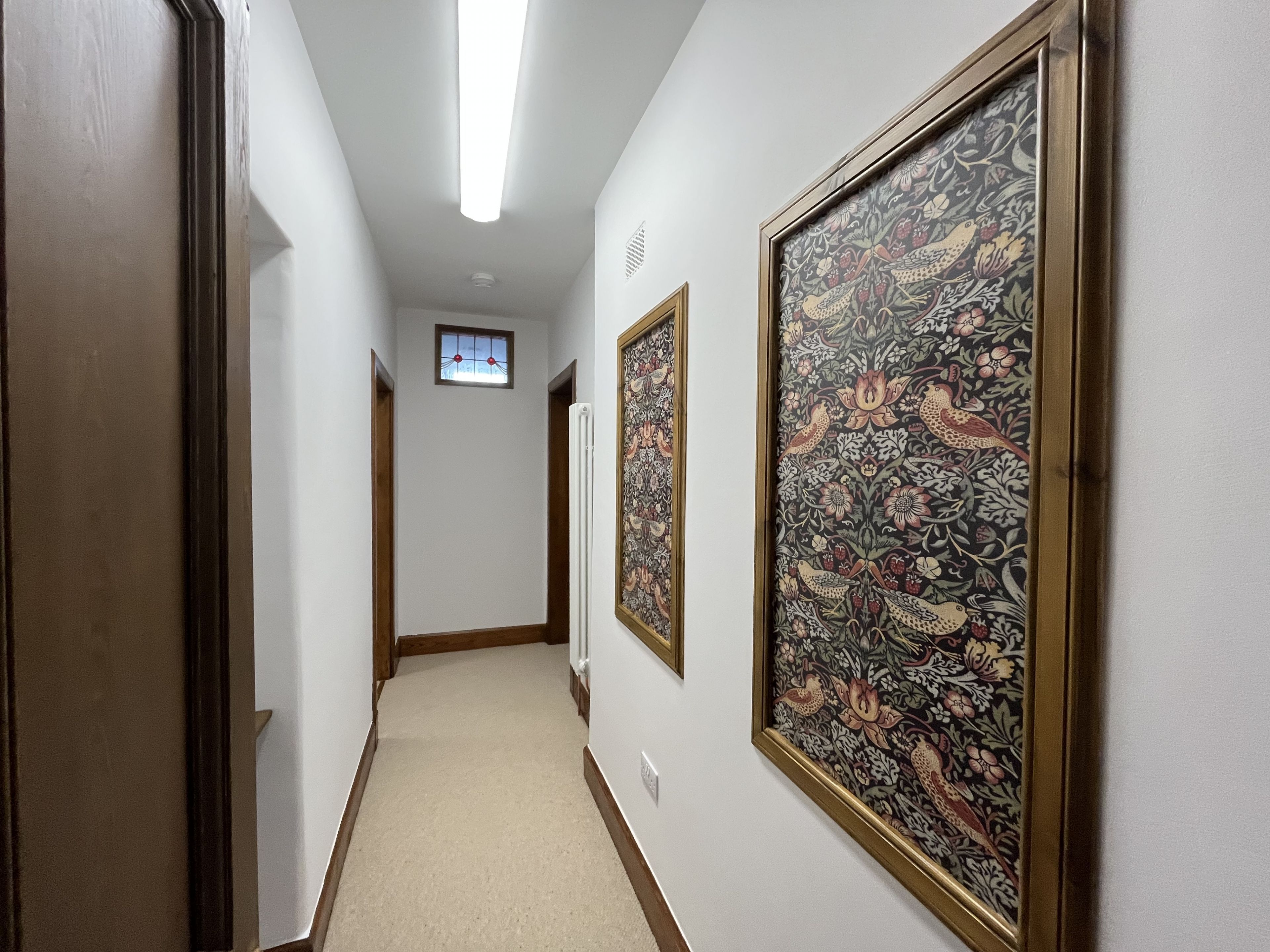 Un couloir aux murs blancs, une porte en bois sur la gauche et deux grands tableaux à motifs sur la droite.