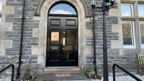 A stone building with a black double door, two black street lamps, and a small bench to the right.