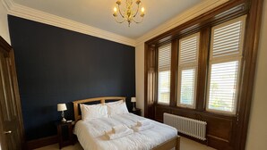 A bedroom with a large bed, two bedside tables, a chandelier, and a window with blinds.