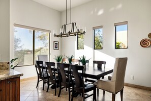 A dining area with a large table, six chairs, and a chandelier. There are potted plants on the table and a view of the outdoors through large windows.