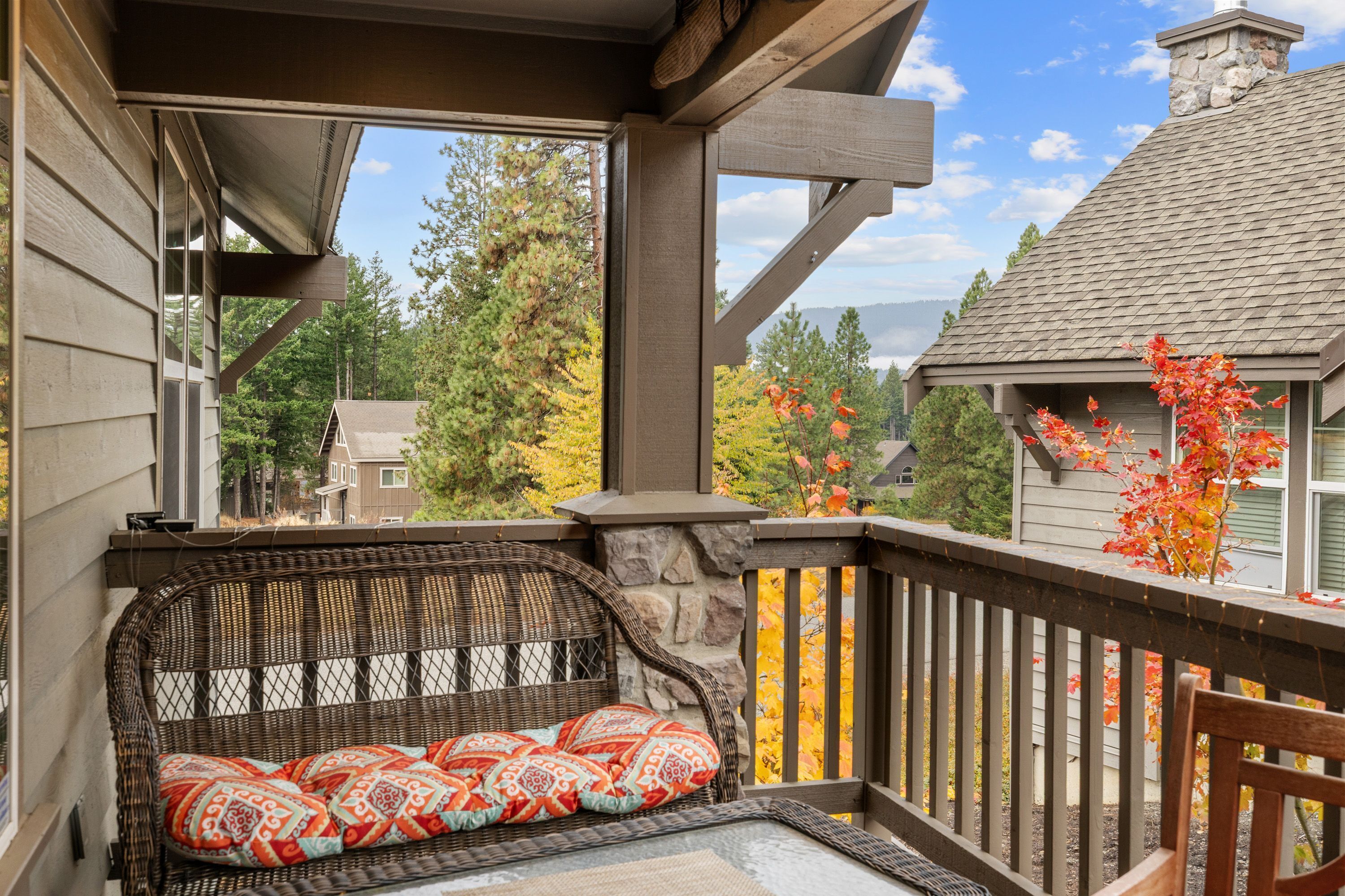 A covered outdoor seating area with a wicker bench and cushions, a stone wall, and a view of surrounding houses and trees.