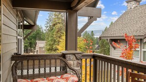 A covered outdoor seating area with a wicker bench and cushions, a stone wall, and a view of surrounding houses and trees. - Raintree at Roslyn Ridge - Cozy Retreat (Ronald)