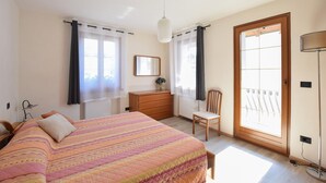 Une chambre à coucher avec un lit, une commode, une chaise et une porte-fenêtre donnant sur un balcon.