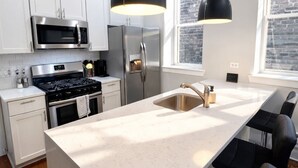 Una cocina moderna con armarios blancos, electrodomésticos de acero inoxidable, una isla central con asientos, iluminación colgante y vistas a una fachada de ladrillo.