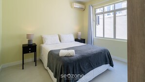 Un dormitorio con una cama, dos mesitas de noche, aire acondicionado y una ventana con cortinas.