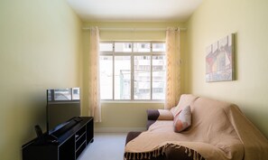 Un salon avec un canapé, un téléviseur posé sur un meuble, une fenêtre avec des rideaux et un tableau accroché au mur.