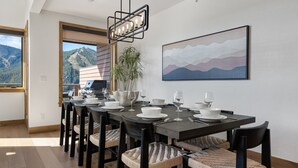 Un comedor con una mesa larga dispuesta para seis personas, rodeado de sillas de mimbre, una lámpara colgante moderna y vistas a la montaña a través de amplios ventanales.