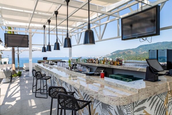 Um bar no terraço com balcão de mármore, banquetas pretas, luminárias pendentes, uma TV de tela plana e vista para o mar e montanhas.