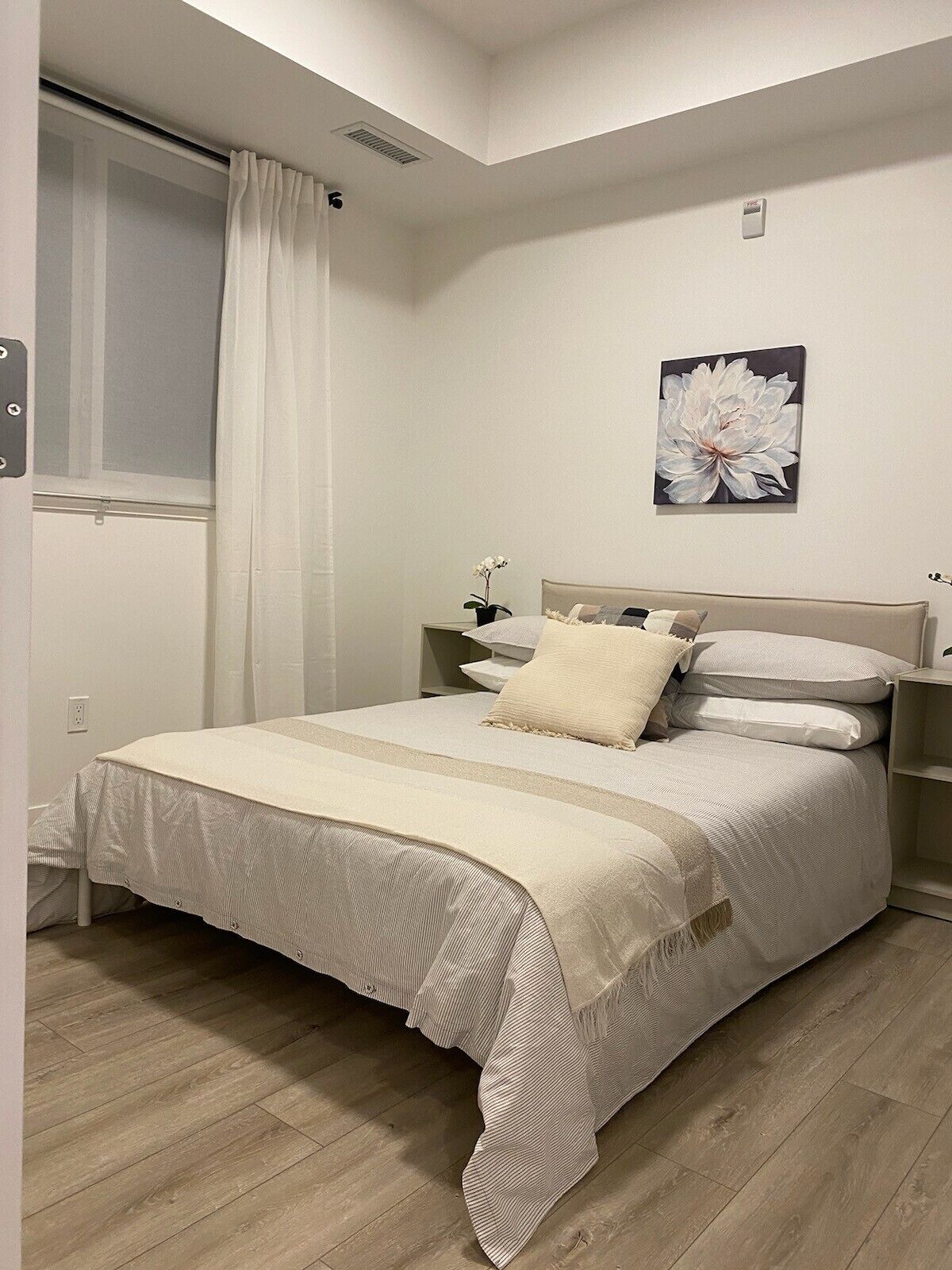 Une chambre à coucher comprenant un lit, une table de chevet, une fenêtre avec des rideaux et un luminaire fixé au mur.