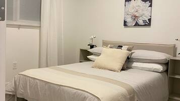 Une chambre à coucher comprenant un lit, une table de chevet, une fenêtre avec des rideaux et un luminaire fixé au mur.