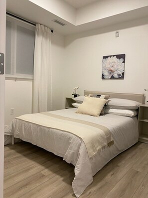 Quarto com cama, mesa de cabeceira, janela com cortinas e luminária fixada na parede.