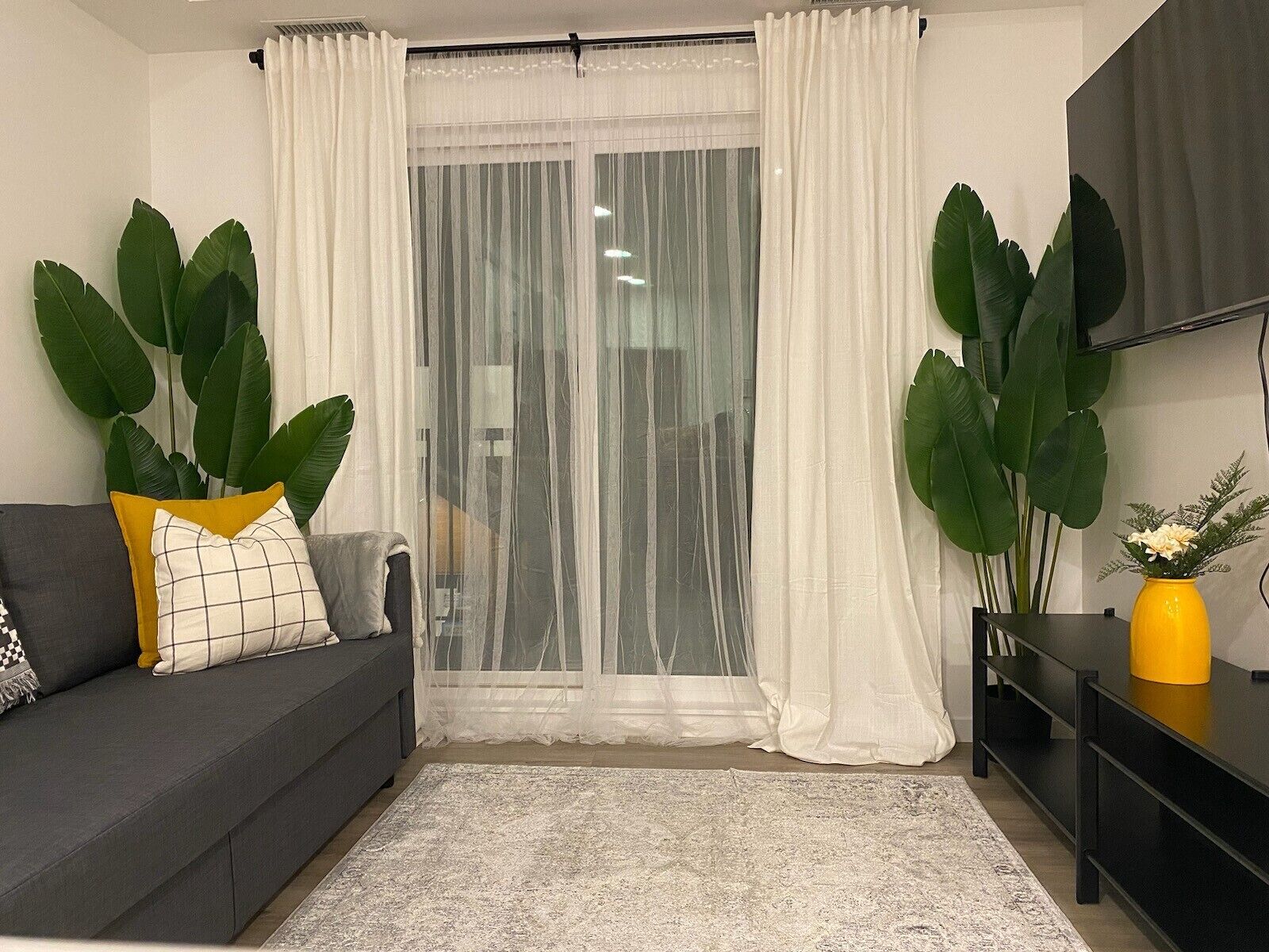 Un salon avec un canapé gris, un vase jaune et de grandes plantes vertes.
