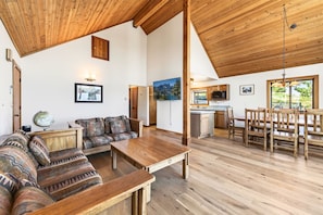 A spacious living area with a wooden ceiling, a sofa set, a dining table, and a kitchen area with wooden cabinets and a view of the outdoors.