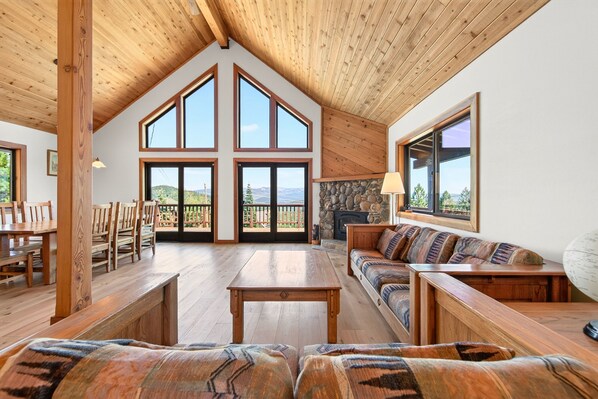 A spacious living area with a high ceiling, wooden beams, large windows, a stone fireplace, and a view of the outdoors.