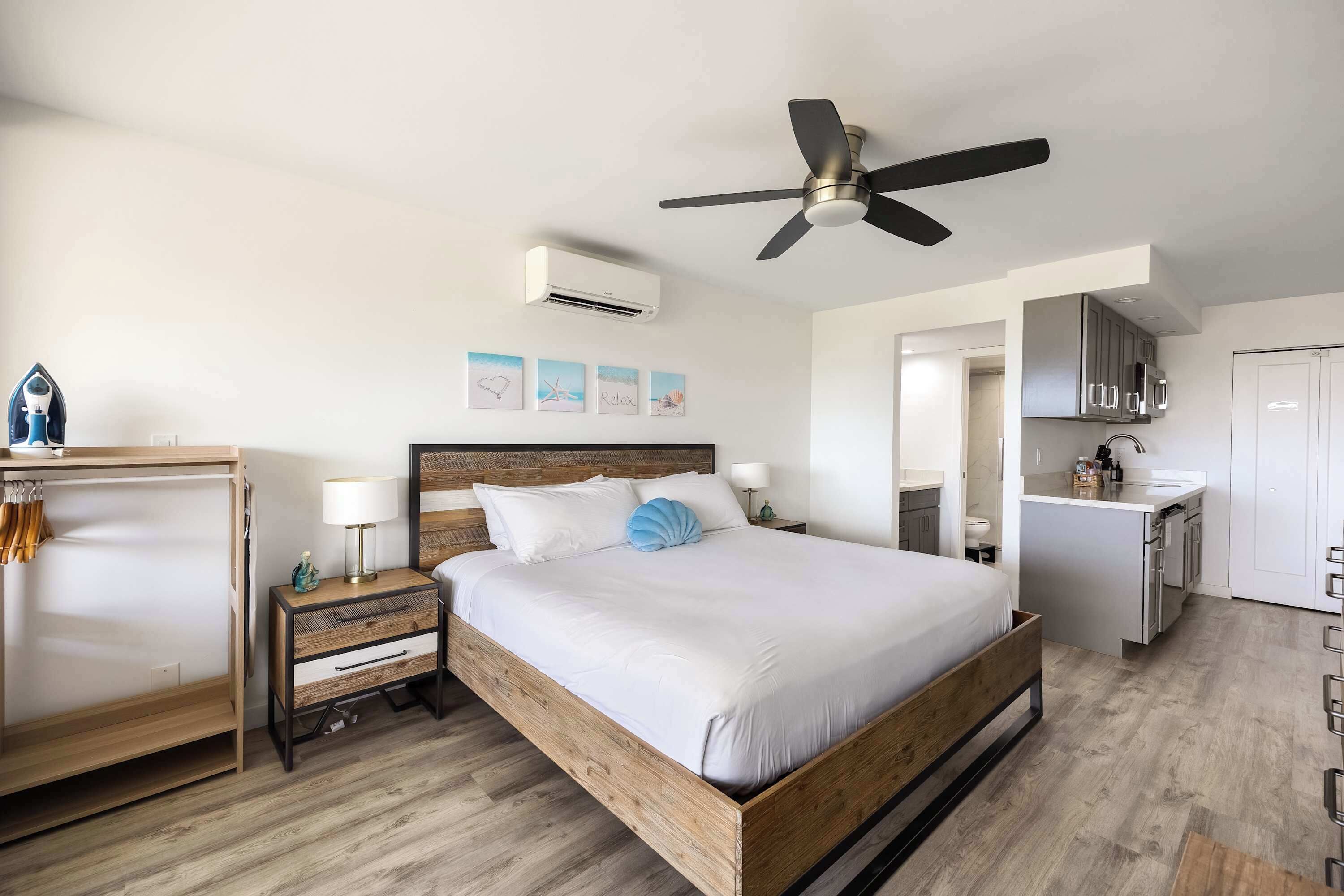 Un dormitorio con una cama de madera, ventilador de techo, mesita de noche con lámpara y una kitchenette al fondo.