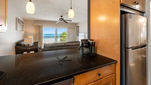 A kitchen with a black countertop, stainless steel refrigerator, wooden cabinets, a ceiling fan, and a view of the ocean through a large window. - Sunset Soul at Pelican Cove Unit 8 by SeaBreeze Vacation (Bradenton Beach)