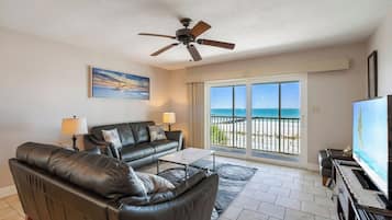 Ein Wohnzimmer mit Sofa, Couchtisch und Fernseher. Es gibt einen Deckenventilator und ein Fenster mit Blick auf den Strand.