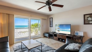 A living room with a large TV, a ceiling fan, a sofa, a coffee table, and a view of the beach through the sliding glass door. - Sunset Soul at Pelican Cove Unit 8 by SeaBreeze Vacation (Bradenton Beach)