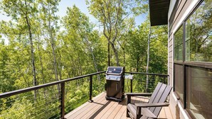 Een houten terras met een barbecue en stoelen, met uitzicht op een bebost gebied.